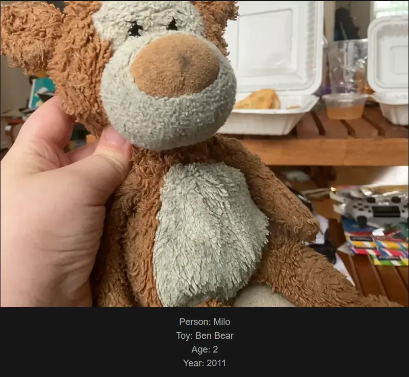 A close-up photo shows a hand holding a well-loved, brown and grey plush teddy bear with a worn, fuzzy texture. In the…