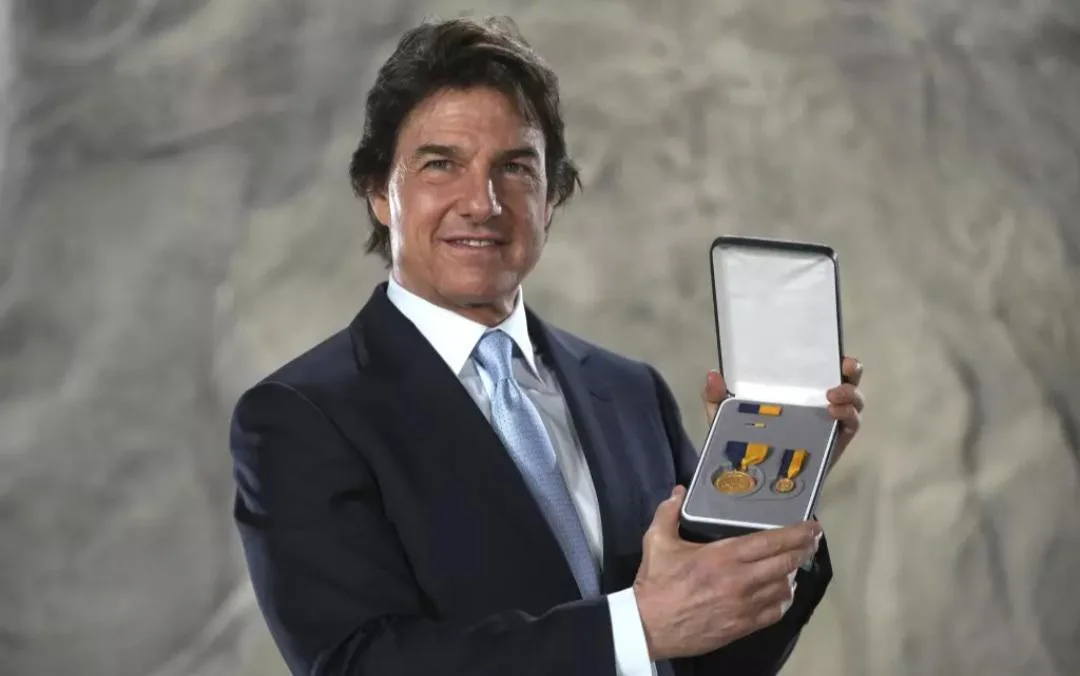 A man in a dark suit and light blue tie holds an open presentation box containing two gold medals with yellow ribbons. He…