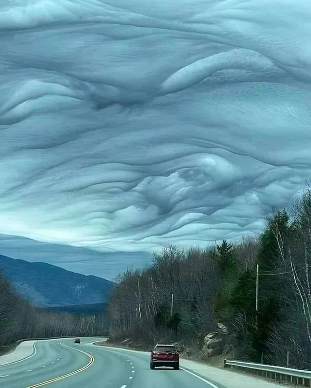 A winding road through a forested mountain area under an unusually textured, wave-like cloud formation that resembles ocean…
