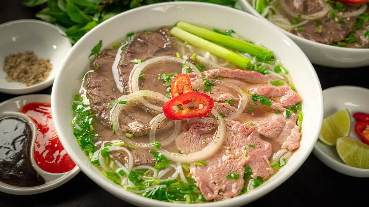 a close-up photograph of a steaming bowl of pho, a traditional vietnamese noodle soup, filled with rice noodles, thinly…