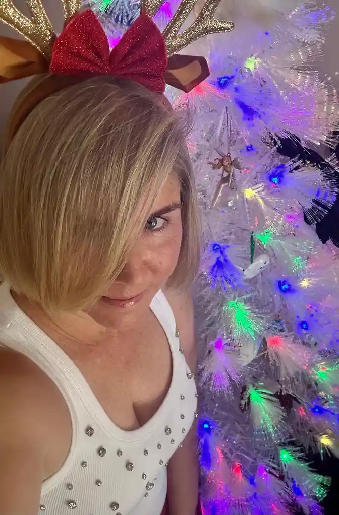 A woman with blonde hair and a white tank top adorned with rhinestones poses in front of a brightly lit, multi-colored…