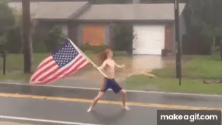 A shirtless person with long hair runs down a wet street while holding an American flag, which flaps wildly in the wind. The…