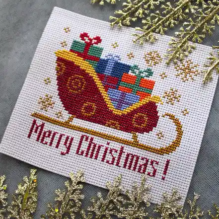 A close-up of a handcrafted cross-stitch Christmas card featuring a red sleigh overflowing with colorful wrapped presents…