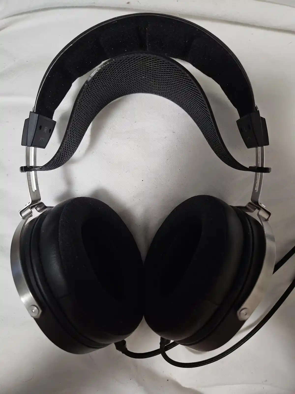 A pair of black over-ear headphones with silver accents rests on a white fabric surface, showing the headband and earcups…