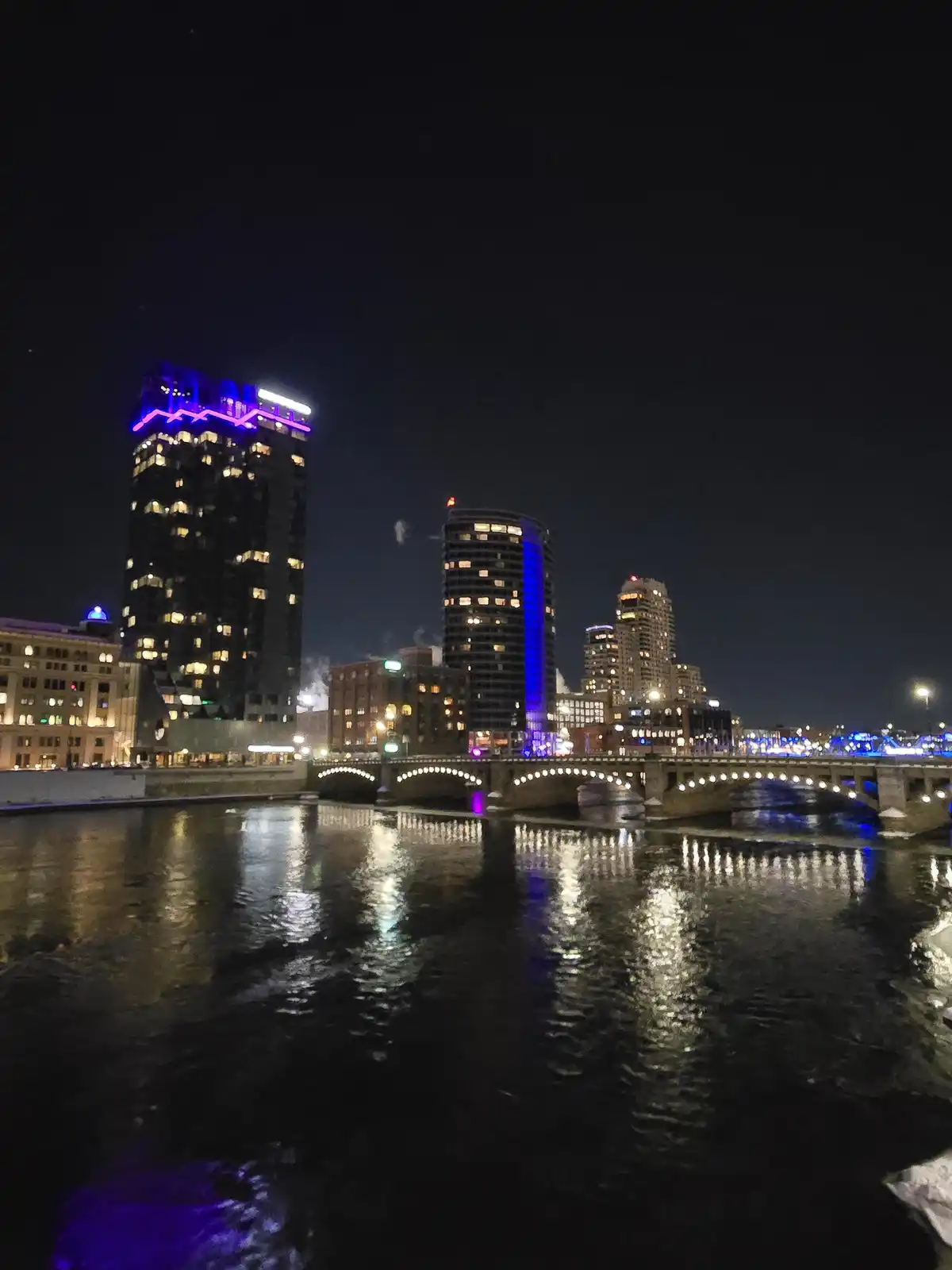 A nighttime cityscape featuring a river with a lit bridge and tall buildings with colorful neon lights reflecting on the…