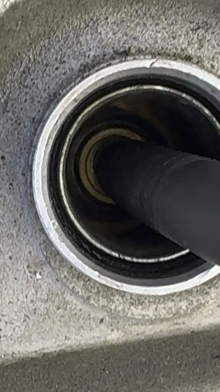 A close-up photo shows a black hose inserted into a circular metal port on a textured gray surface, likely a vehicle's fuel…