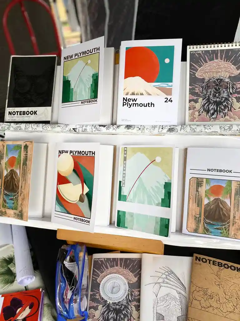 A shelf displays various notebooks with artistic covers, including some labeled 'New Plymouth' with mountain illustrations…