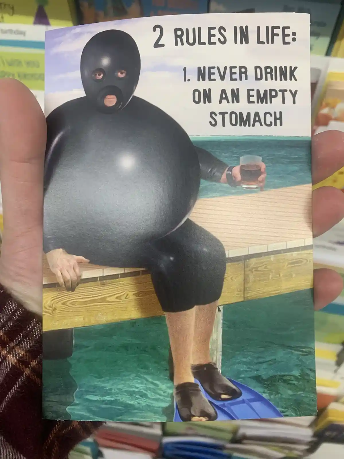 A person wearing a black, full-body wetsuit and a black mask with eye and mouth holes is sitting on a wooden dock by the…