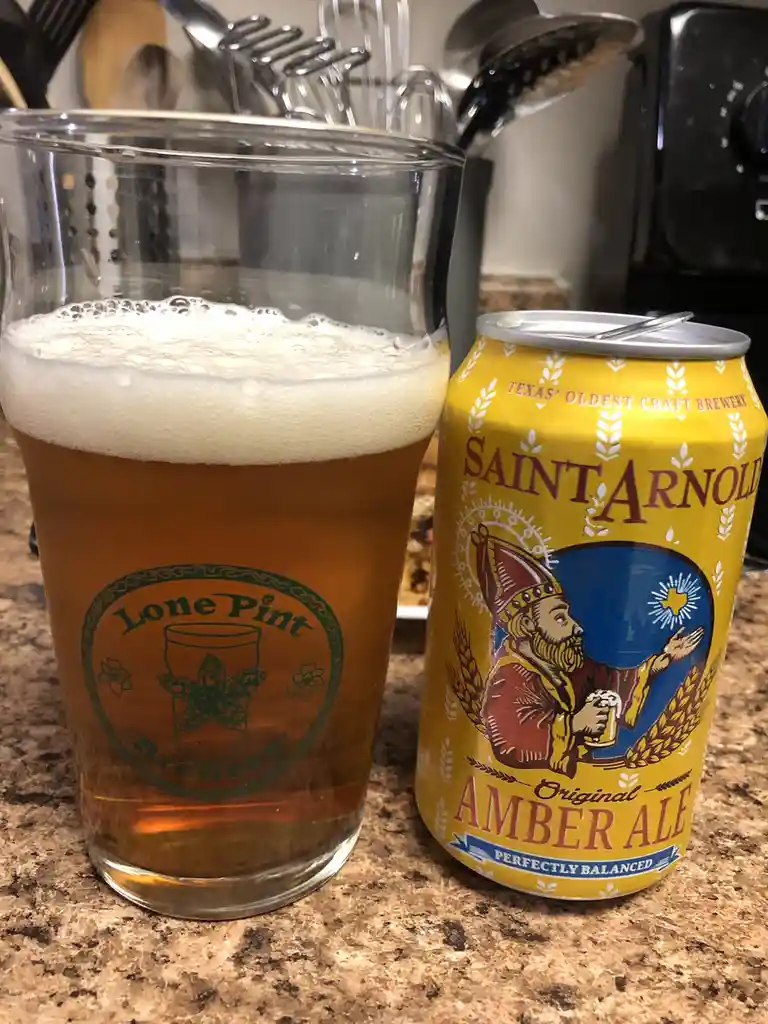 A can of Saint Arnold Original Amber Ale sits next to a glass of beer on a kitchen counter, the can's label featuring a…