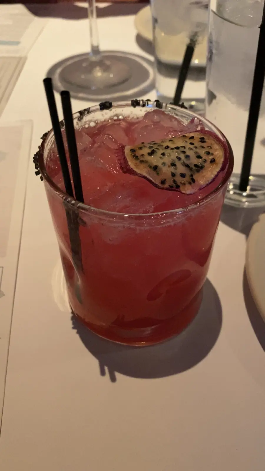 A close-up photo of a vibrant pink cocktail in a rocks glass, garnished with a slice of dragon fruit and two black straws…