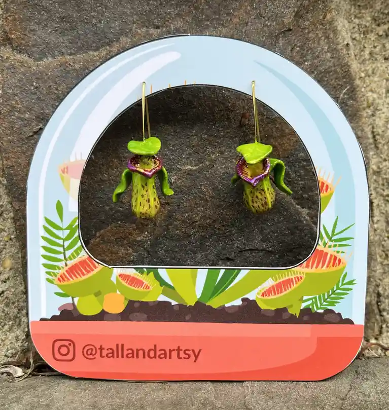A pair of small, green pitcher plant-shaped earrings with purple rims hang from gold hooks inside a decorative, arched…