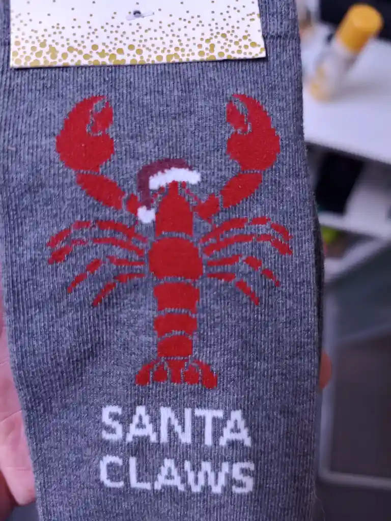 A close-up of a gray sock featuring a red knitted lobster wearing a Santa hat, with the text 'SANTA CLAWS' knitted below it…