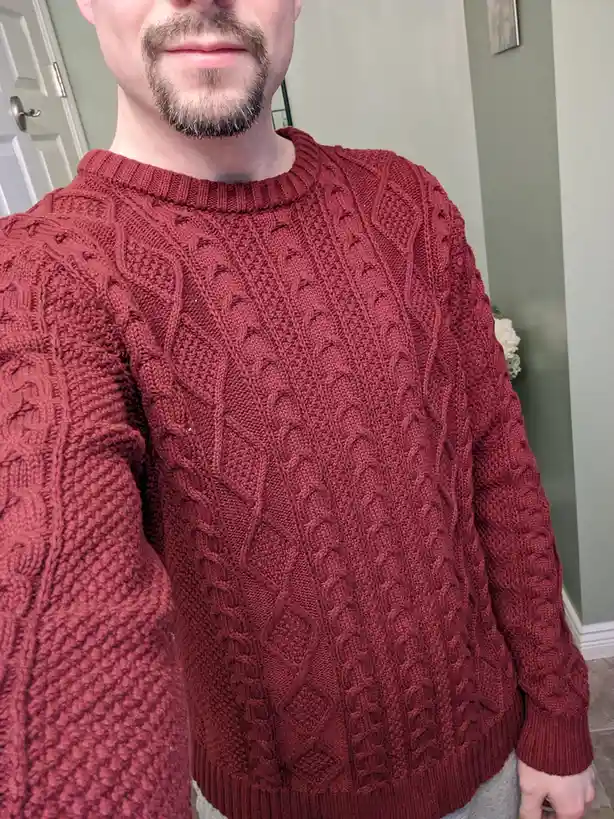 A man with a goatee is shown from the chest up, wearing a red cable-knit sweater. The sweater has a prominent textured…