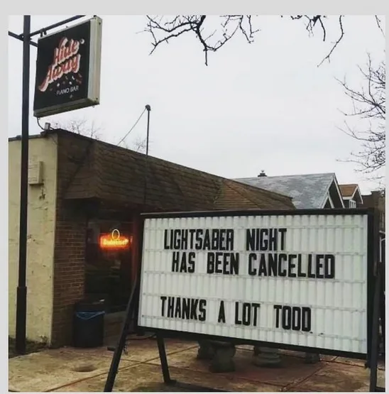 An outdoor sign in front of a bar named 'Hide Away Piano Bar' reads 'LIGHTSABER NIGHT HAS BEEN CANCELLED THANKS A LOT TODD'…