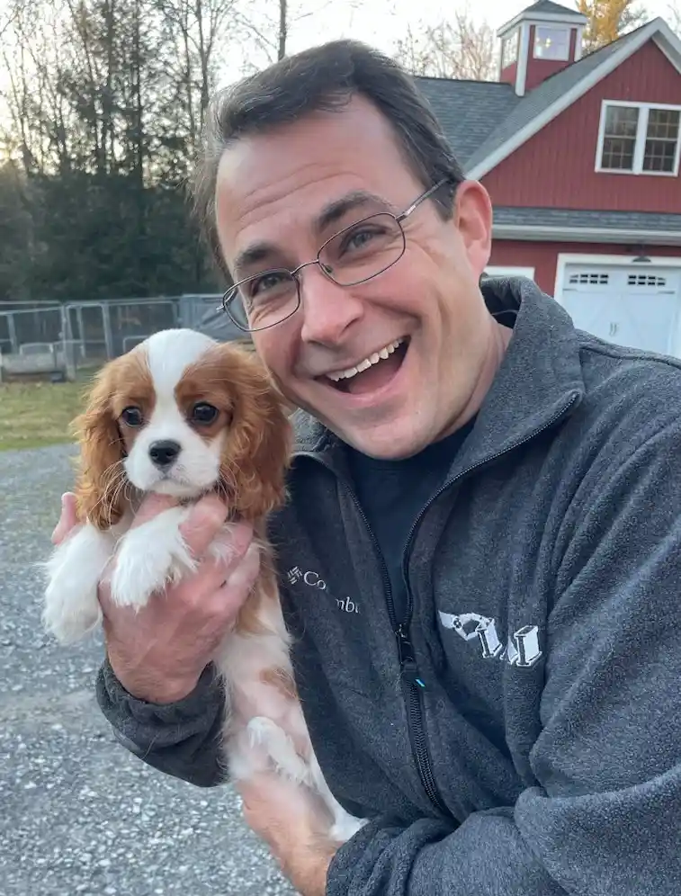 A man with glasses and a grey Columbia jacket is holding a small brown and white Cavalier King Charles Spaniel puppy. He is…
