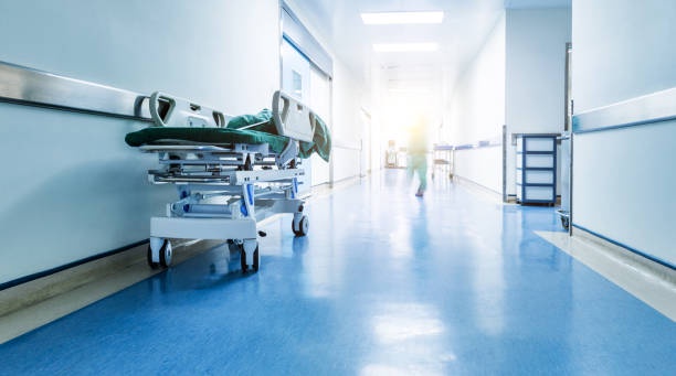 A hospital hallway with a gurney parked on the left side, a person in scrubs walking away down the brightly lit corridor…