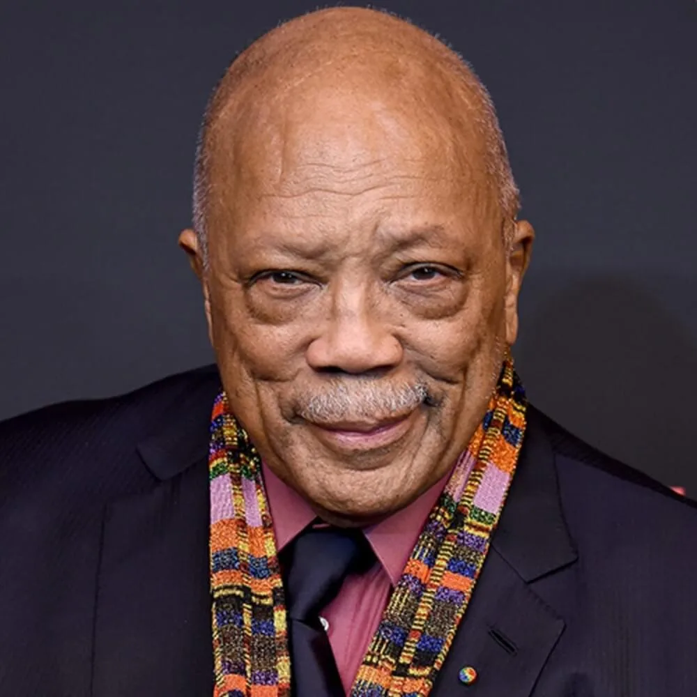 A close-up photo of an older Black man with a bald head and mustache, wearing a dark suit, pink shirt, dark tie, and a…