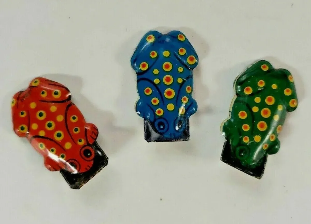Three small, colorful, hand-painted frog-shaped objects lie side by side on a plain white surface. Each frog is a different…