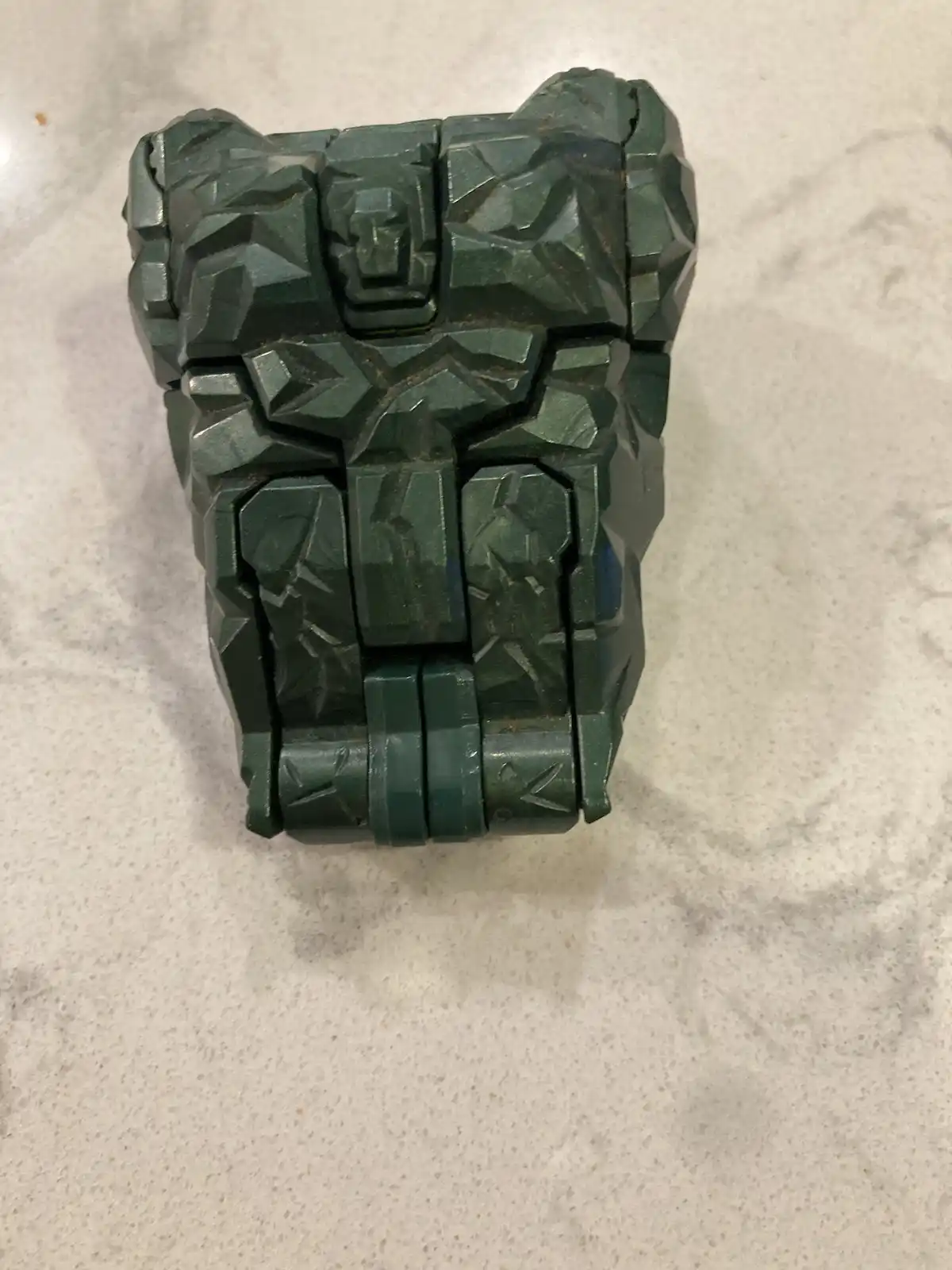 A dark green, angular, and heavily textured object resembling a robot or armor torso sits on a light speckled countertop…