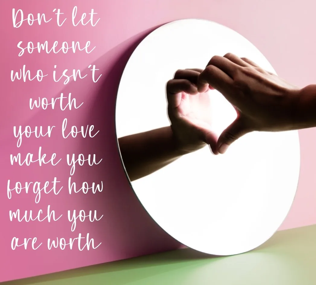 A pair of hands forms a heart shape in front of a circular mirror against a pink background, with white cursive text…