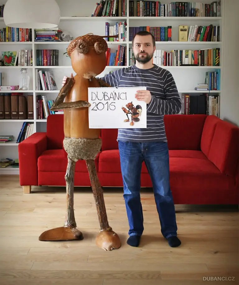 A man in a striped shirt and jeans stands beside a tall, wooden, stick-figure-like sculpture with a round head and twig…