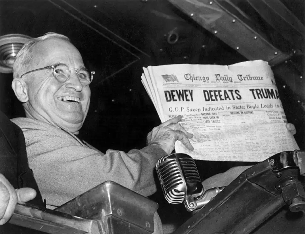 A black-and-white photo shows a smiling man in glasses holding up a newspaper titled 'Chicago Daily Tribune' with the…