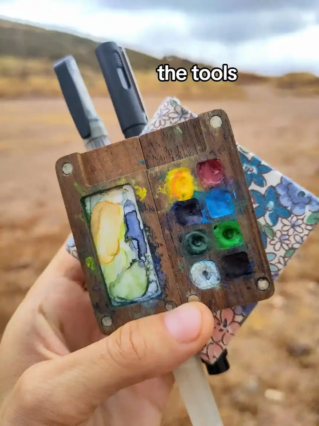 A hand holds a small, weathered wooden artist's palette with dried paint blobs in various colors, attached to a patterned…