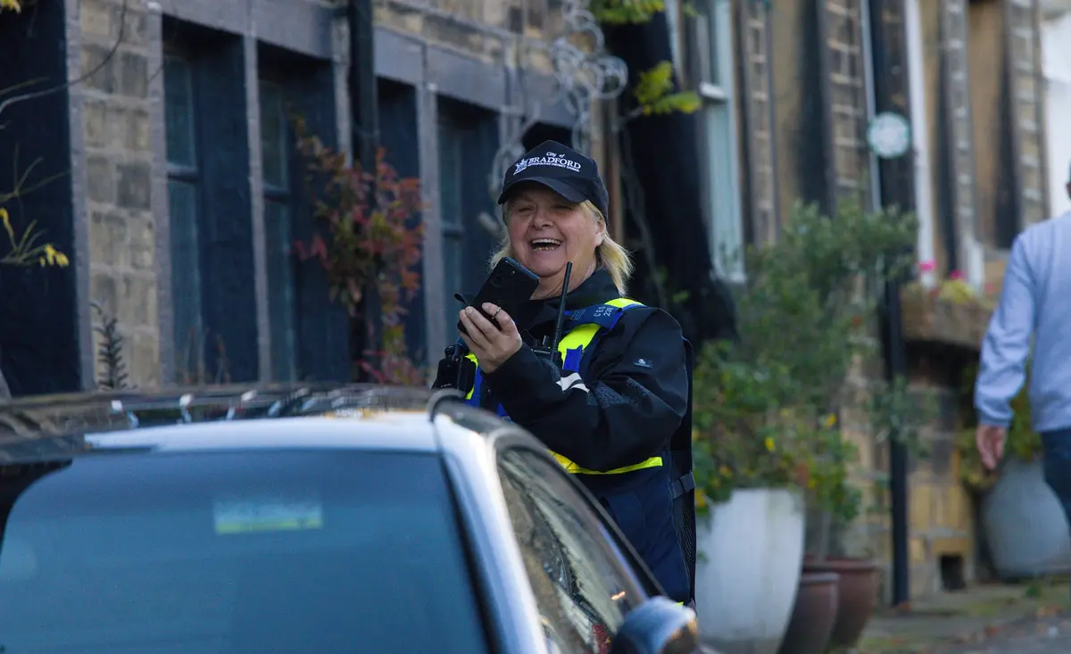 A woman in a Bradford City Council uniform, smiling broadly while holding a smartphone, stands next to a car on a…