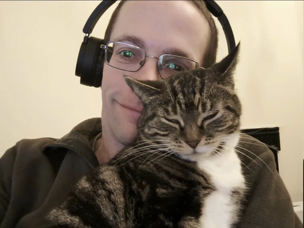 A man wearing black over-ear headphones and glasses smiles while holding a tabby cat with a white chest. The cat has its…