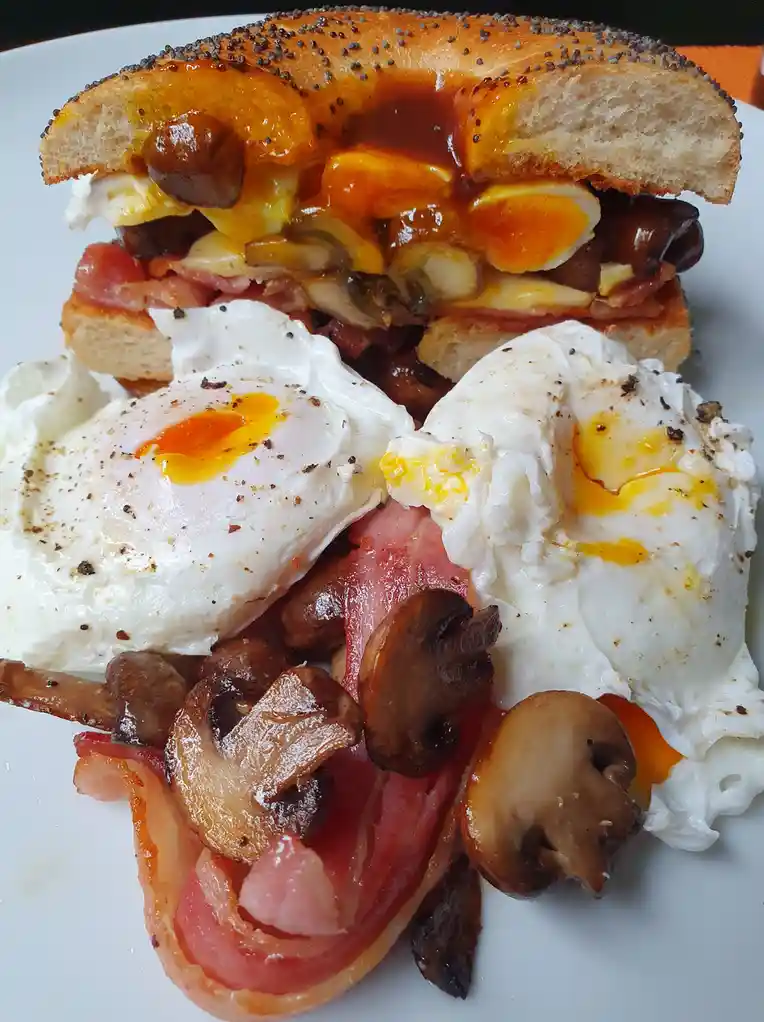 A plate of breakfast food featuring two poached eggs with runny yolks, bacon, sautéed mushrooms, and a bagel sandwich loaded…