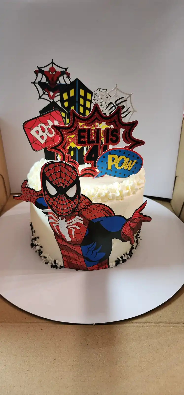 A white frosted birthday cake decorated with Spider-Man themed toppers, including a large Spider-Man figure on the side, a…