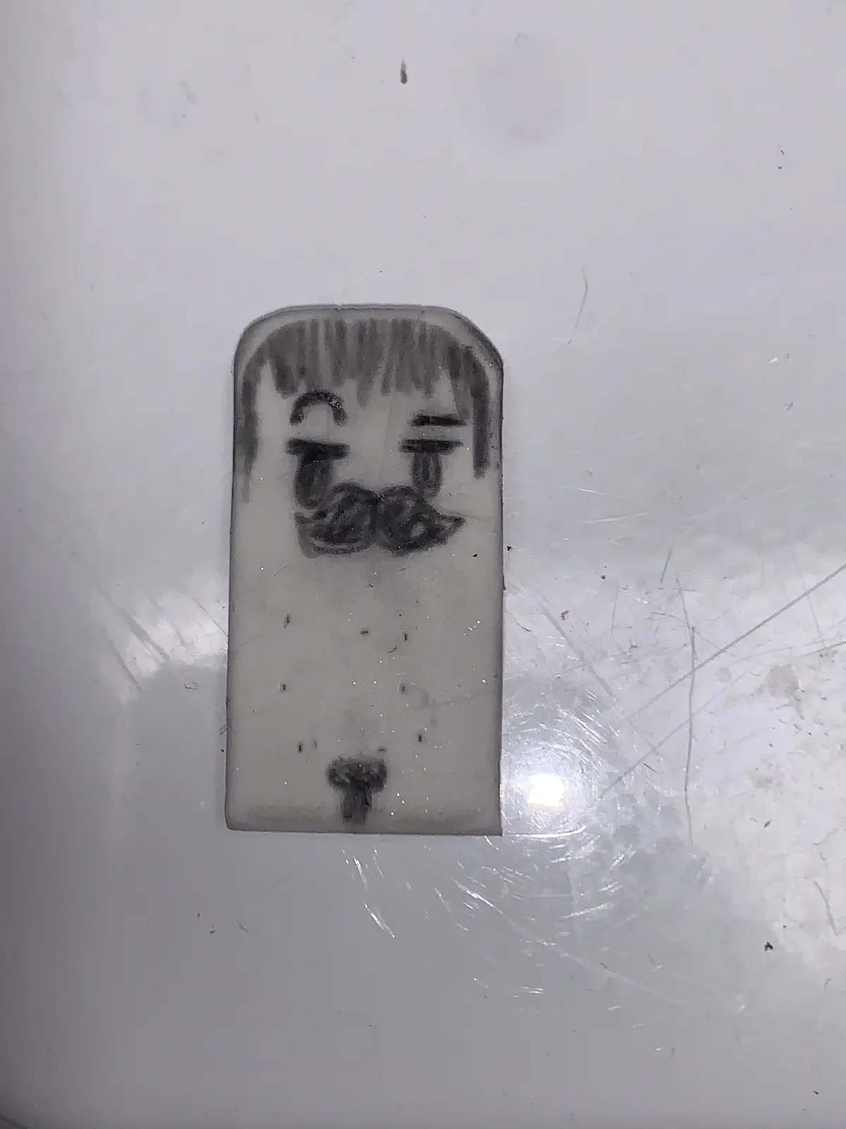 A crudely drawn cartoon face with tears and a mustache is sketched on a rectangular, slightly worn white surface, likely a…