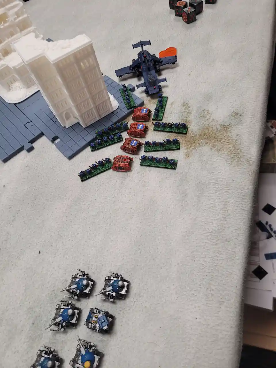 A tabletop wargaming setup shows miniature models of blue and red armored vehicles and infantry units arranged in combat…