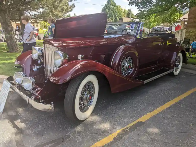 A shiny maroon vintage convertible with its hood open is parked at an outdoor car show, surrounded by people. The overlaid…