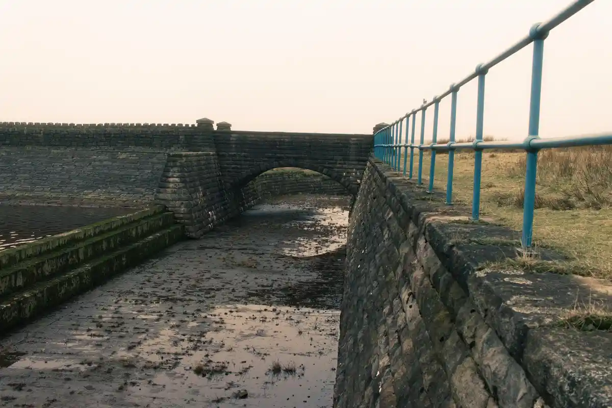 A low-angle photo shows a stone arch bridge over a shallow, muddy canal with mossy steps on the left and a blue metal…