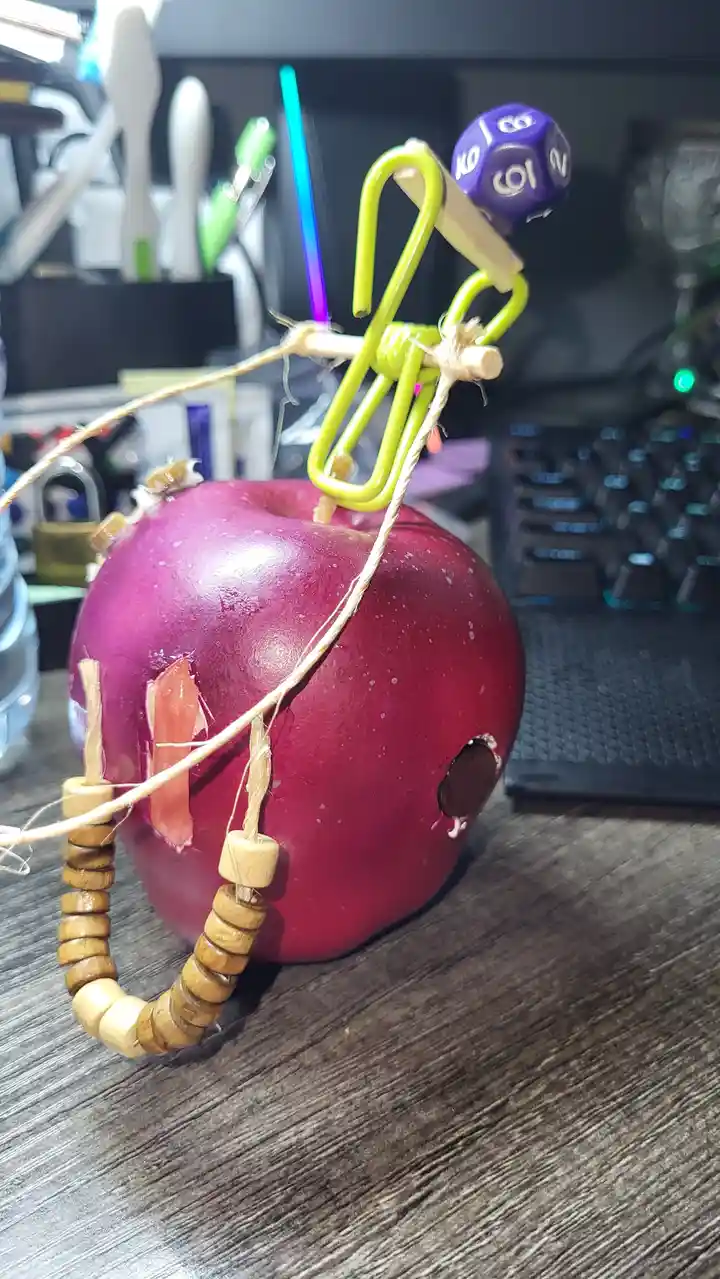A red apple on a desk has been crudely modified with twine, wooden beads, and a green paperclip to resemble a character with…