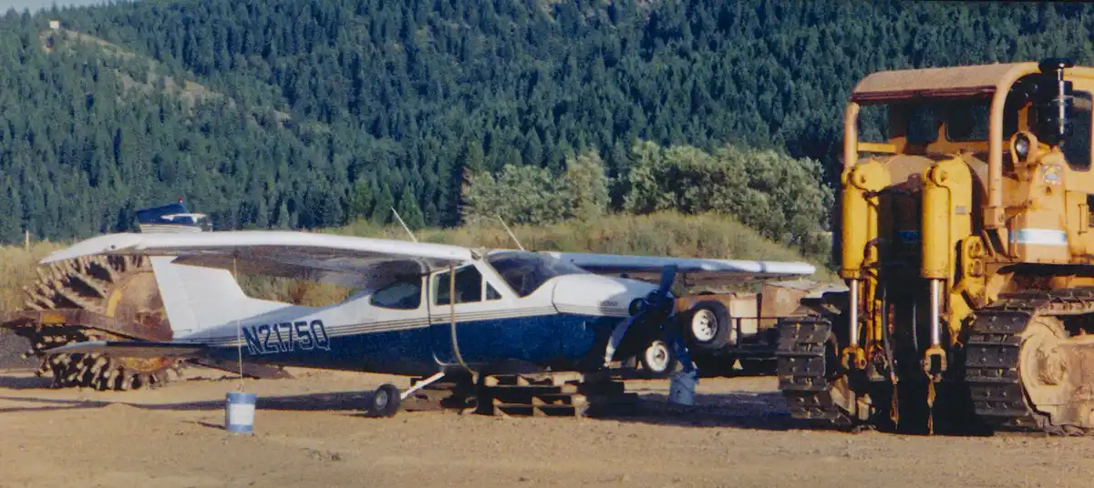 A small blue and white airplane with registration N2175Q is parked on a dirt field next to a large yellow bulldozer. The…