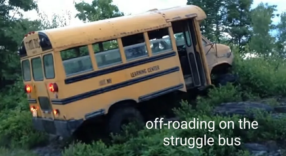 A yellow school bus is tilted and stuck in dense green foliage, its front end angled upward and rear lights illuminated. The…