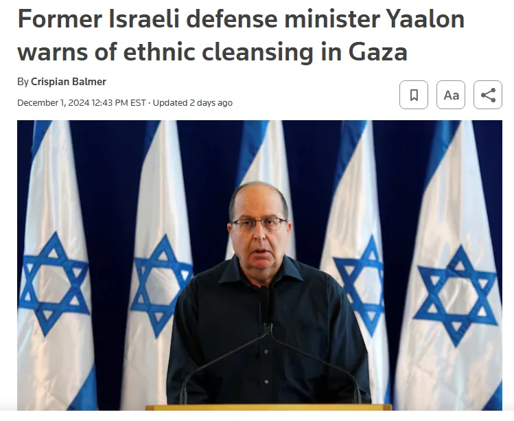 A man in a black shirt and glasses stands at a podium in front of multiple Israeli flags, appearing to speak at a press…