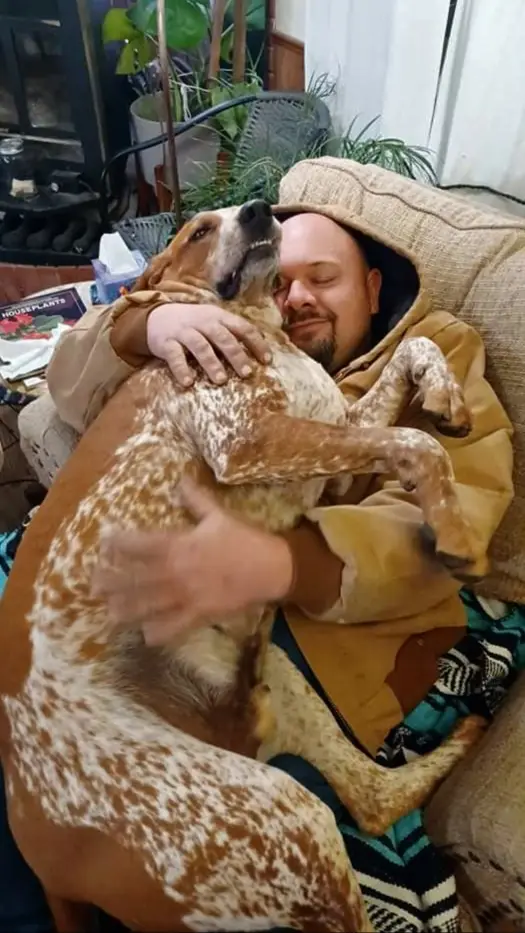 A man in a tan hoodie is cuddling a large, brown-and-white spotted dog on a couch. The dog is lying across his chest with…