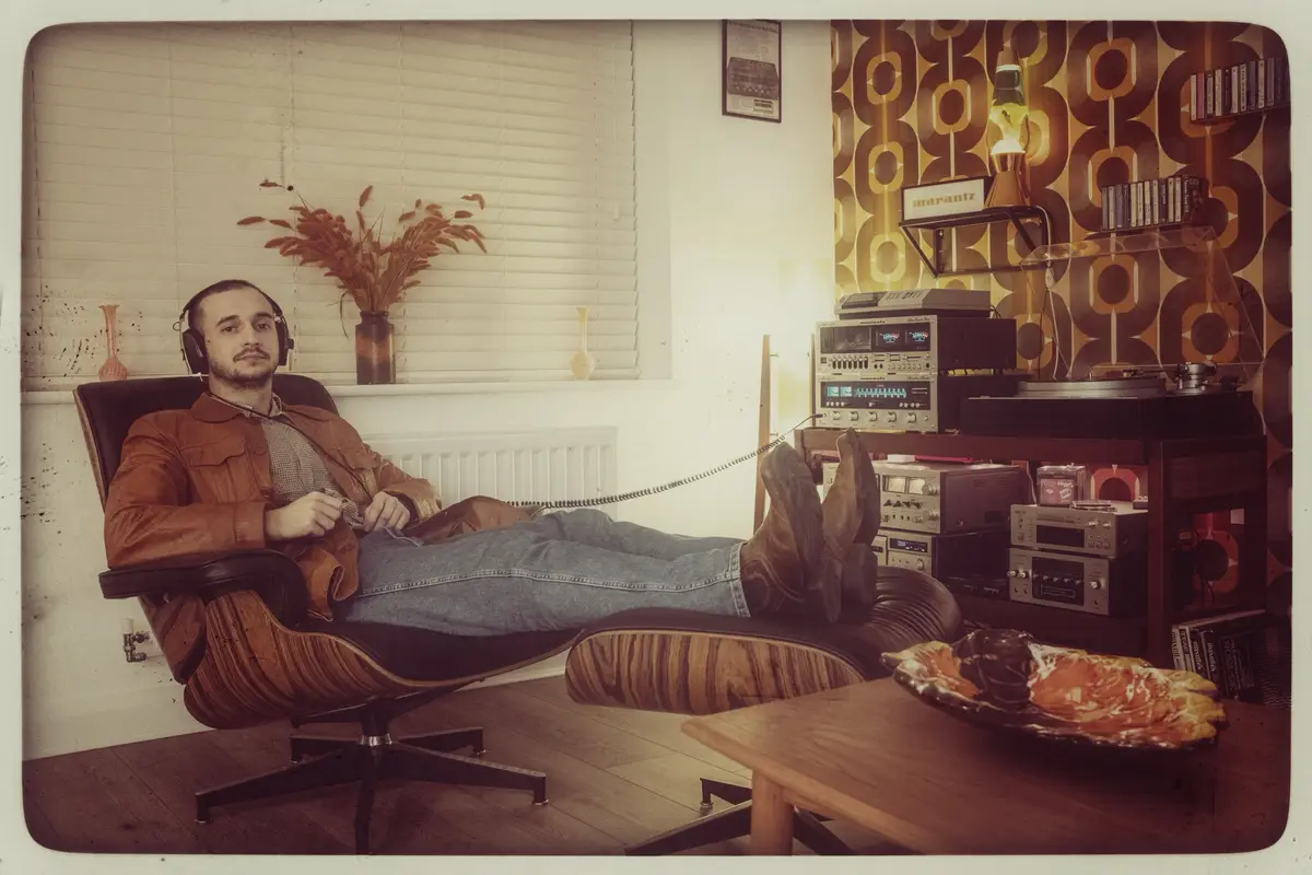 A man with headphones sits relaxed in a wooden lounge chair with his feet up on a matching ottoman, surrounded by vintage…