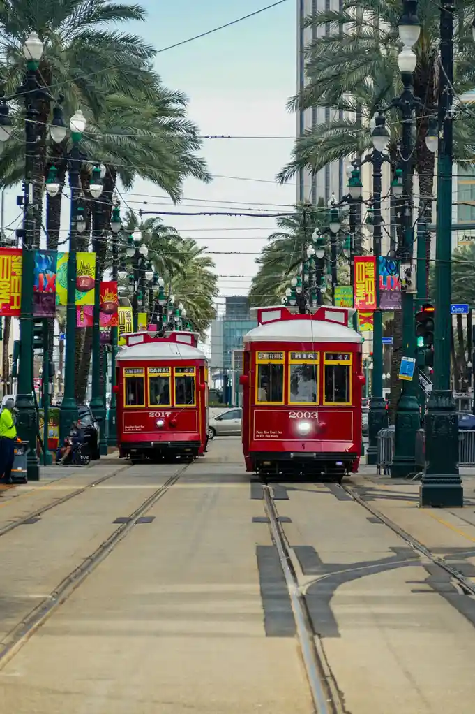 Two red New Orleans streetcars face each other on parallel tracks, one labeled '2017' and the other '2003', with palm trees…