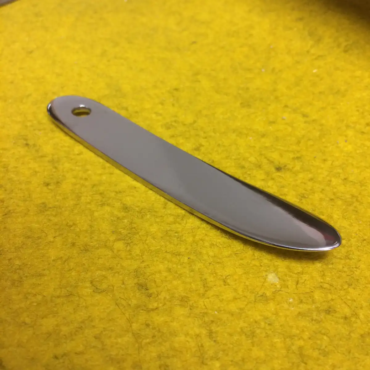 A shiny, silver, elongated metal object with a rounded tip and a hole near one end lies diagonally on a textured yellow…