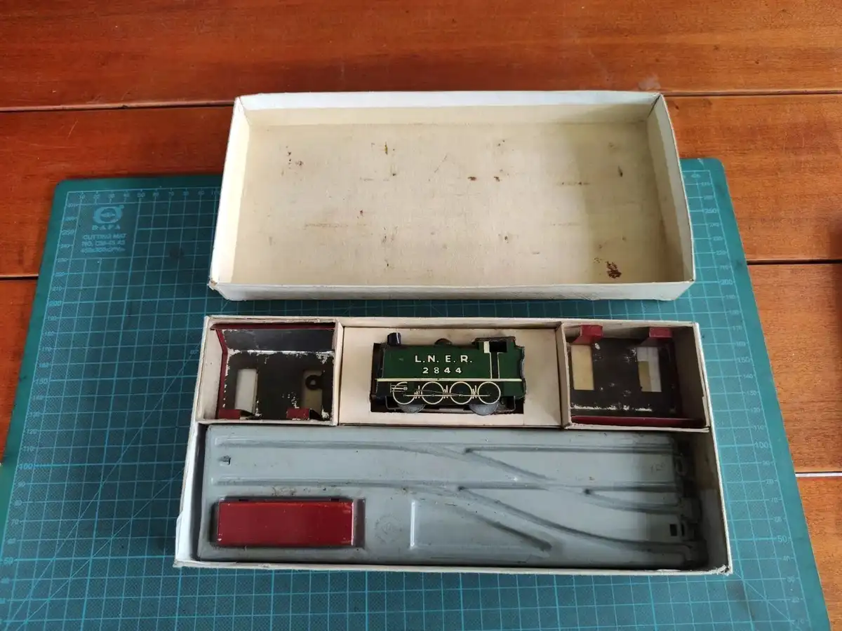 A vintage toy train set is displayed in its open cardboard box on a green cutting mat, showing a green locomotive labeled…