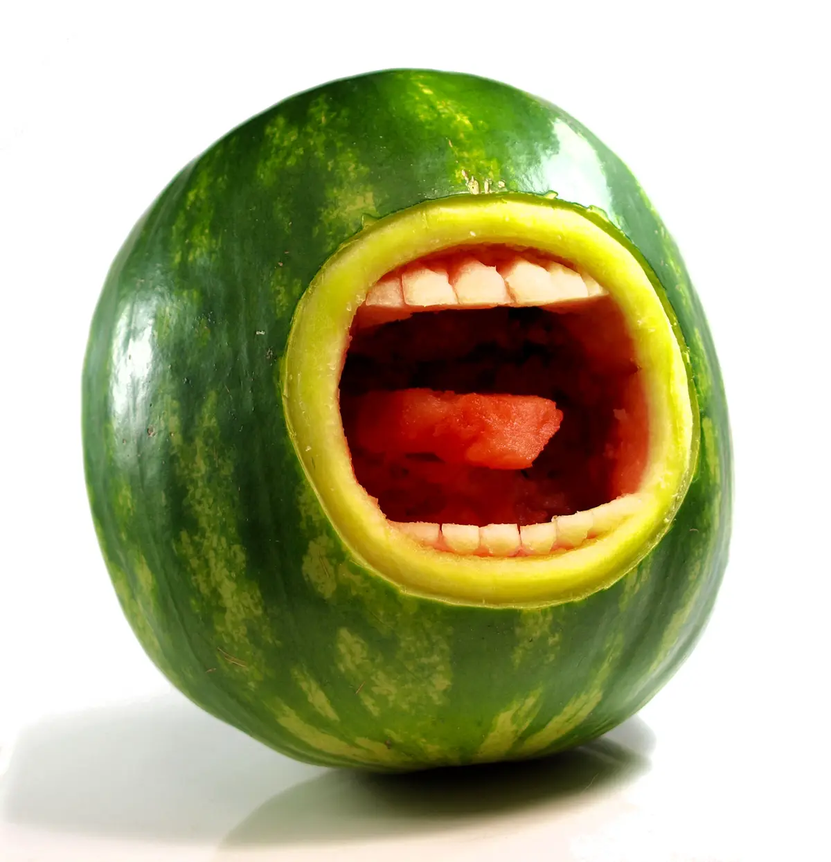 A whole watermelon has been carved to resemble a human face with an open mouth, featuring rows of white rind teeth and red…