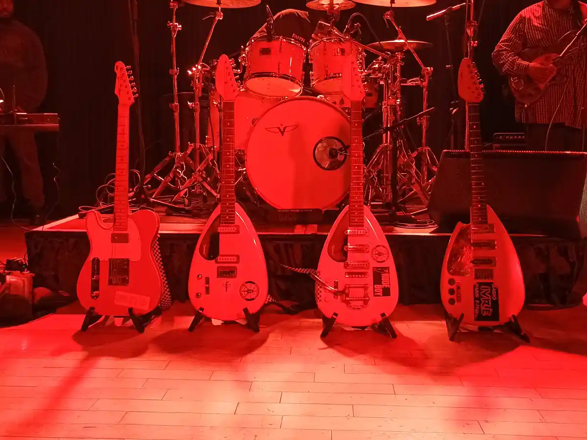 A stage bathed in red light features four electric guitars on stands in front of a drum kit, with musicians partially…
