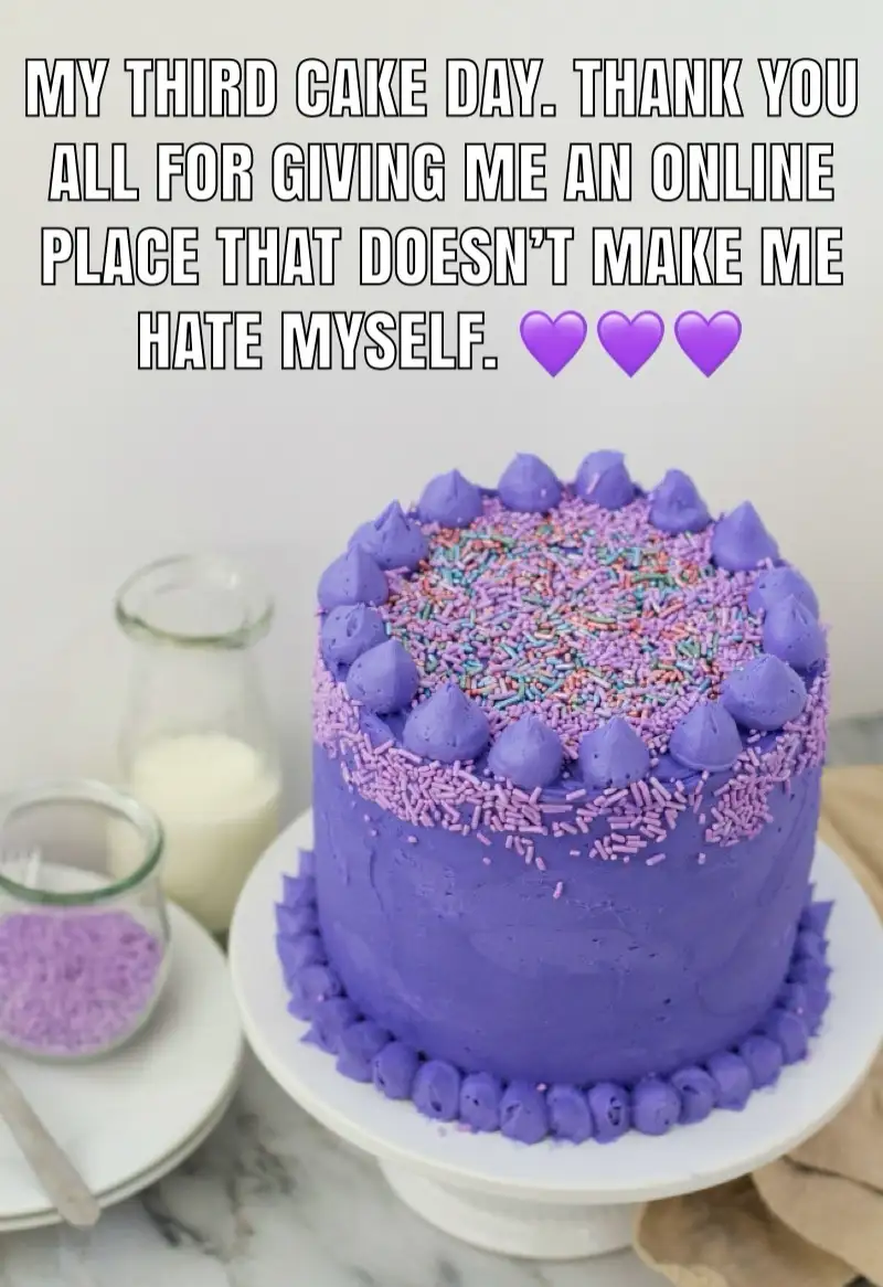 A round purple-frosted cake with colorful sprinkles and piped frosting decorations sits on a white cake stand. In the…