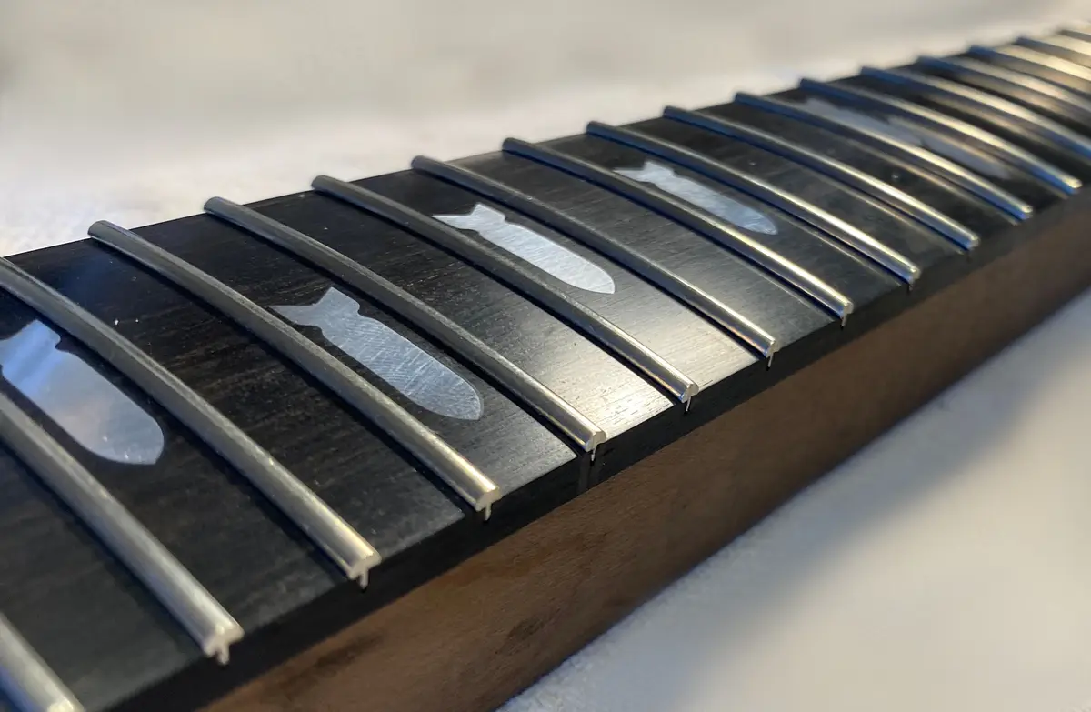 A close-up photo of a guitar fretboard with silver metal frets, where the inlays between the frets are shaped like fish. The…