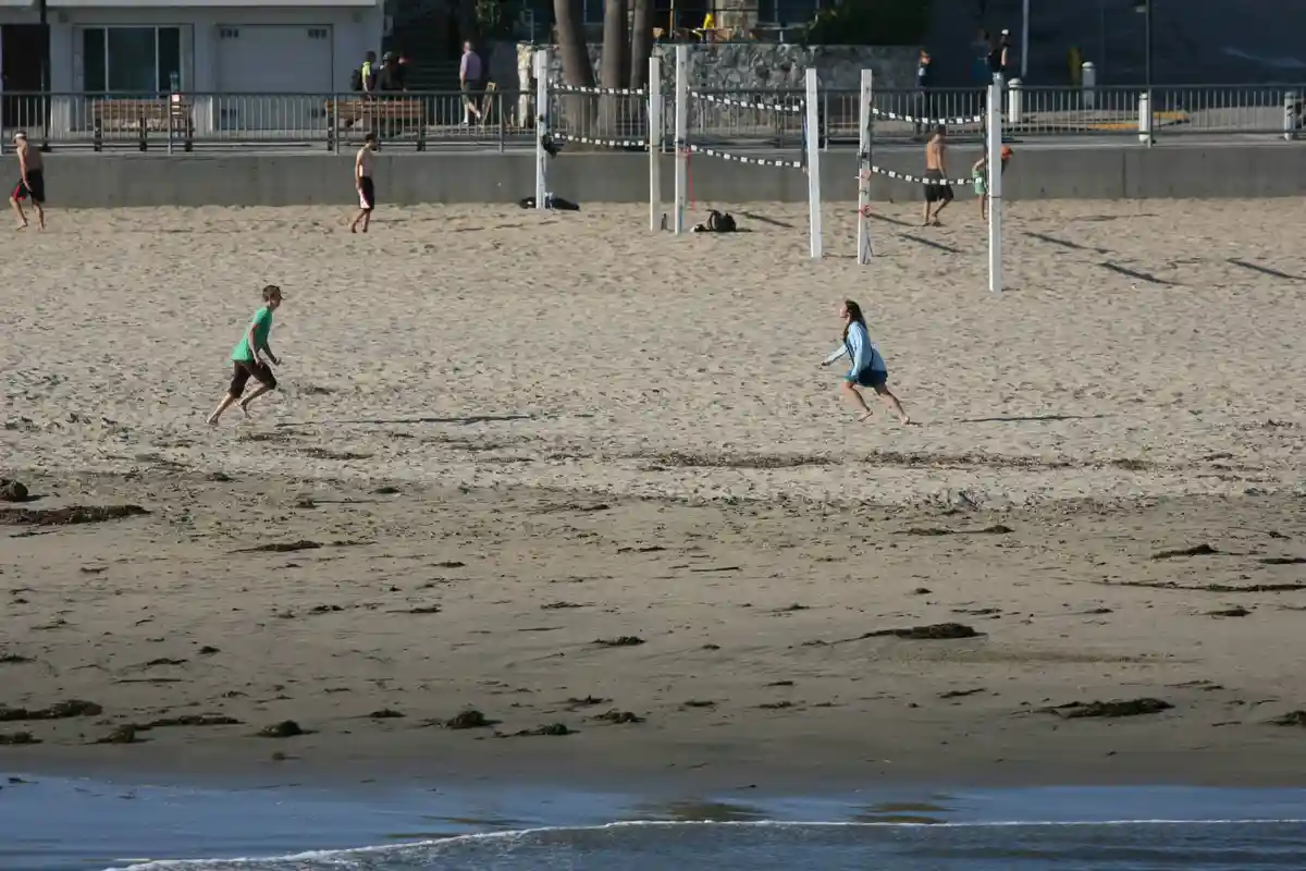 A beach scene with two people running toward each other across sand near the water, with volleyball nets and other…