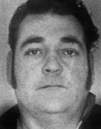 A grainy, black-and-white mugshot-style photo of a man with a serious expression, dark hair, and a collared shirt. Overlaid…
